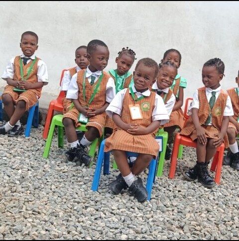 A class of pupils in tan pinafore uniforms and green ties sitting on small chairs on the schoolyard gravel